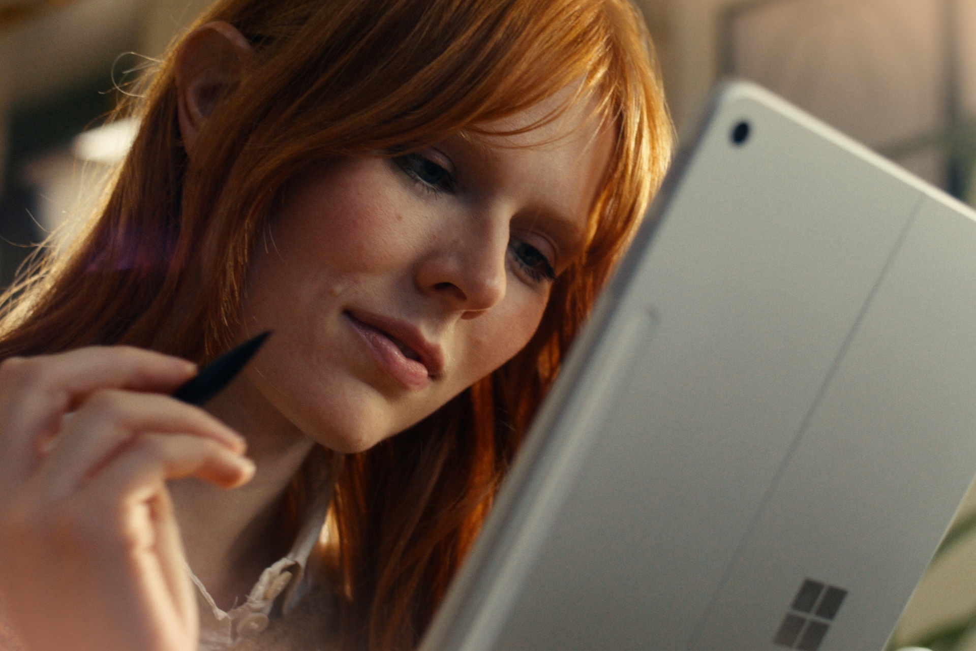A woman with red hair uses a stylus to write or draw on a Microsoft Surface tablet, looking focused and engaged.