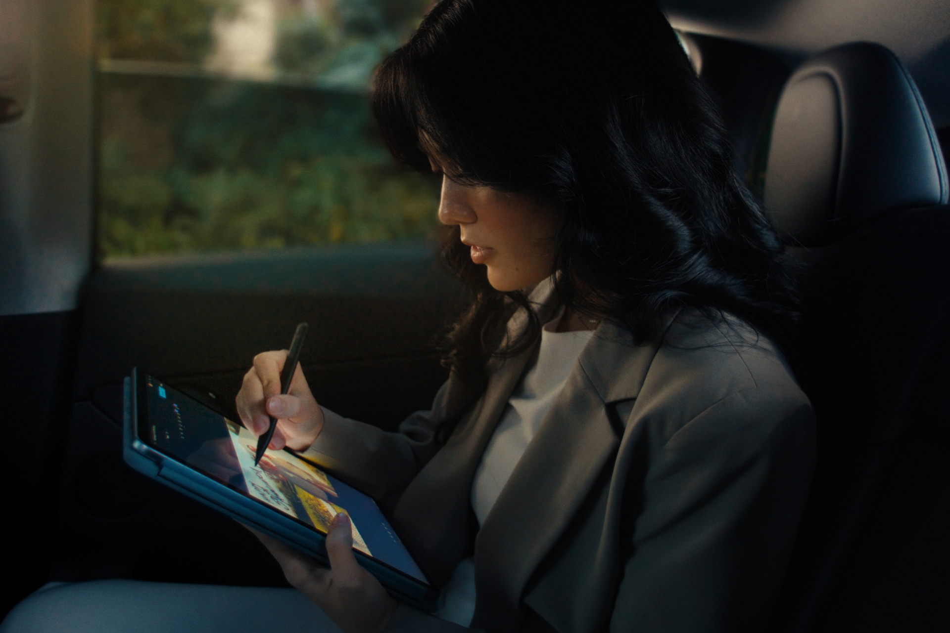 A woman sits in the backseat of a car, using a stylus to draw on a tablet. She wears a light-colored blazer and focuses intently on her work. Natural light filters through the car window.