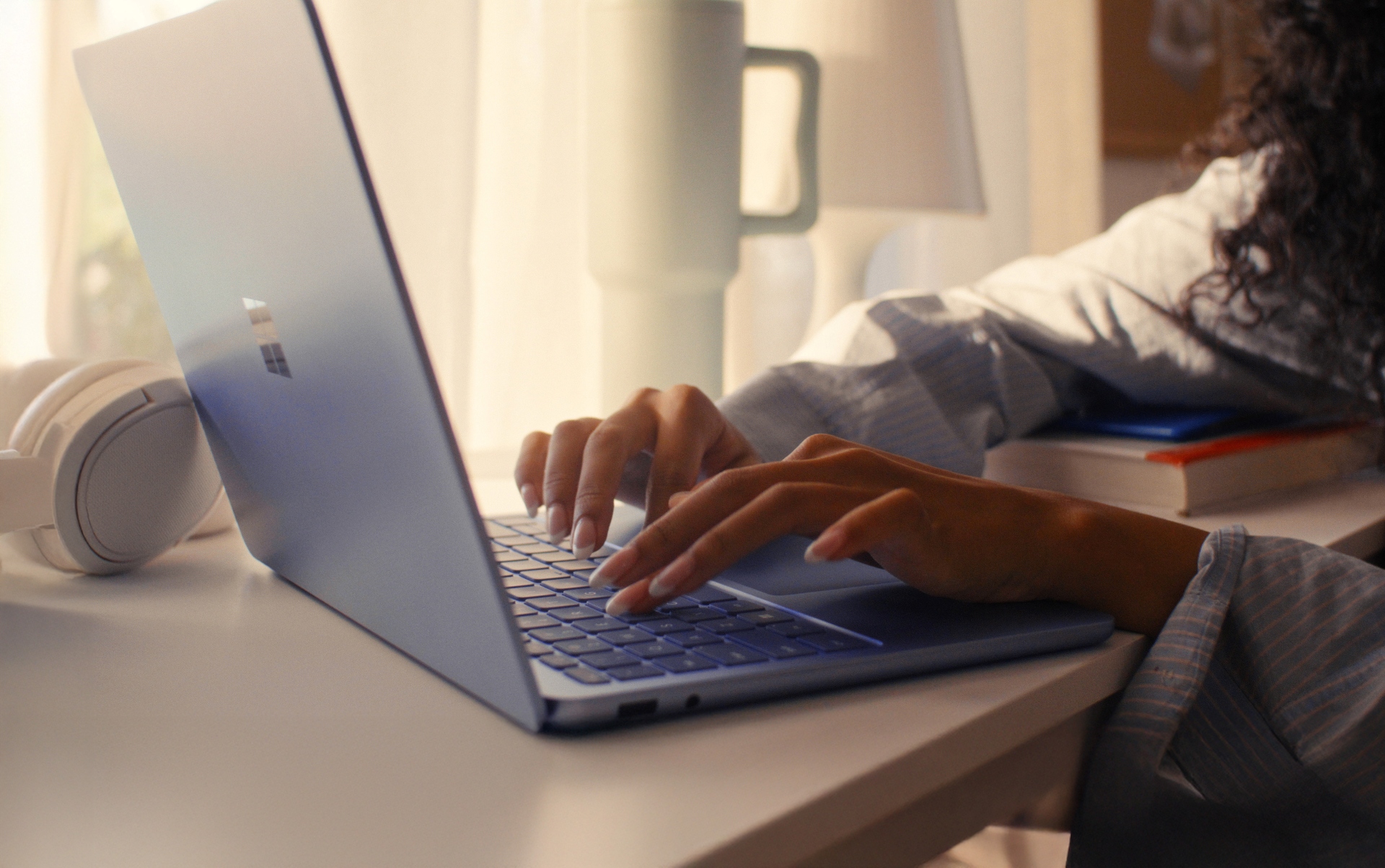 A person types on a blue laptop at a desk, with a stack of books, a lamp, and white headphones nearby, sunlight streaming through a curtain in the background.