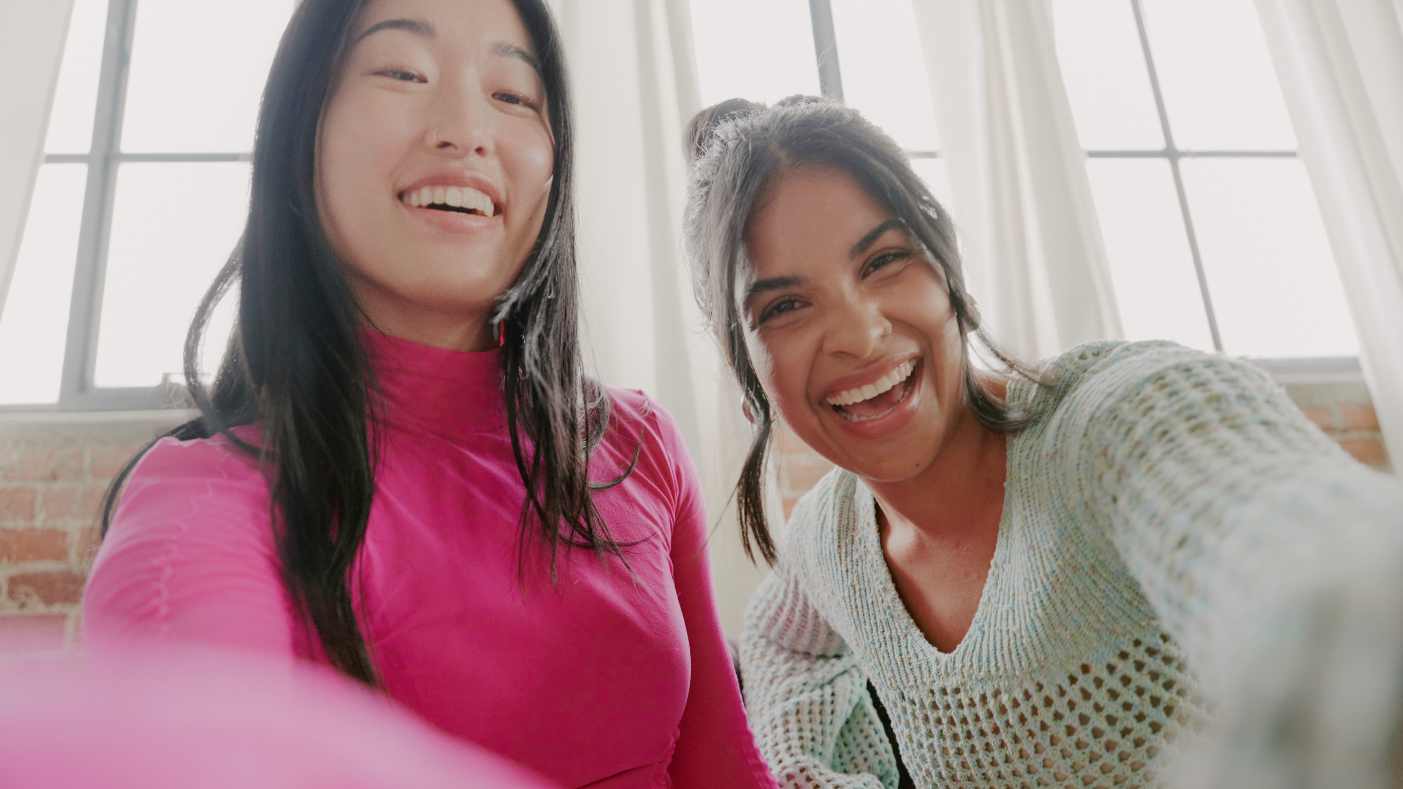 Two women smiling and laughing together while taking a selfie indoors, near large windows with sheer white curtains, just after completing a quick sign-in. One wears a bright pink top, the other a light blue sweater.