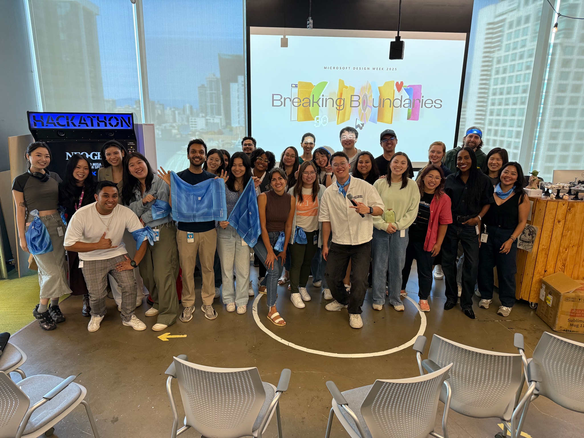A diverse group of people smile and pose together in an office space with “Breaking Boundaries” displayed on a screen behind them. Some hold blue bandanas, and hackathon signage is visible on the left.