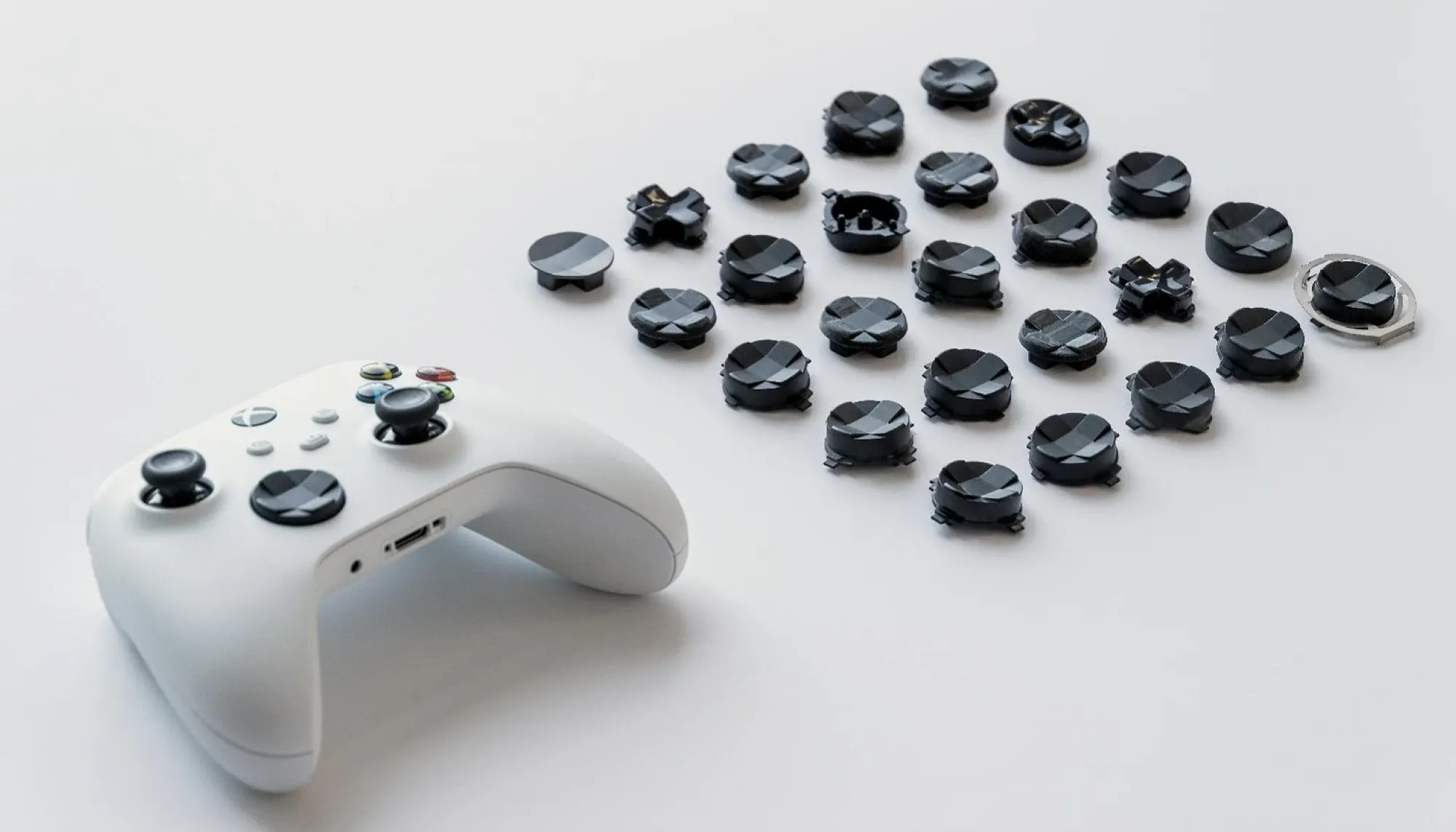 A white gaming controller sits on a white surface next to an organized array of black interchangeable analog stick tops arranged in rows.