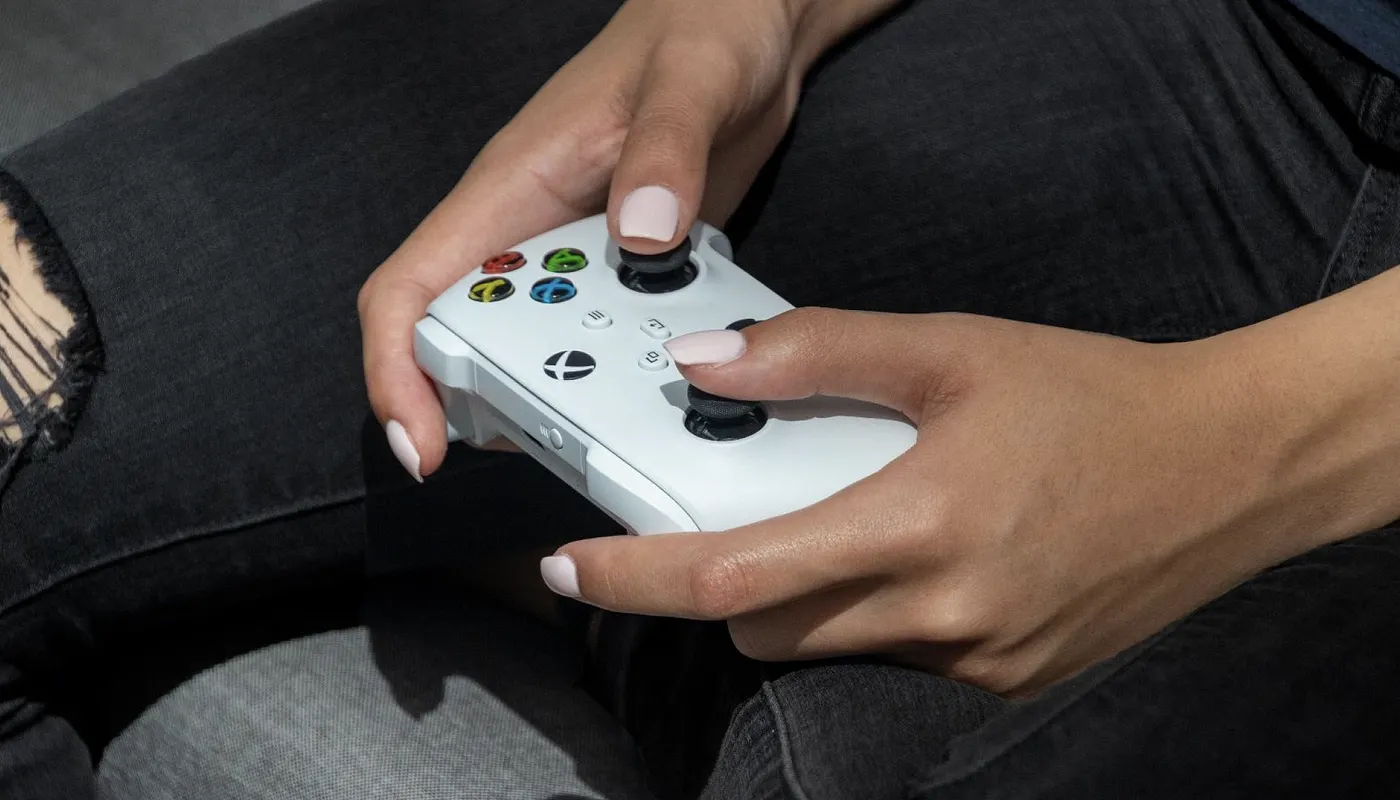 A person with light pink nail polish holds a white Xbox controller while sitting, wearing dark jeans. The focus is on their hands and the controller.
