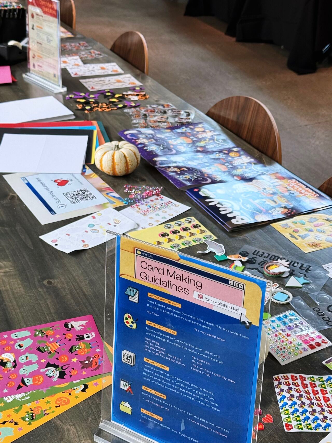 A table set up for a card-making activity with colorful stickers, paper, markers, instruction sheet, and a small decorative pumpkin. Several Halloween-themed items and puzzles are also visible on the table.