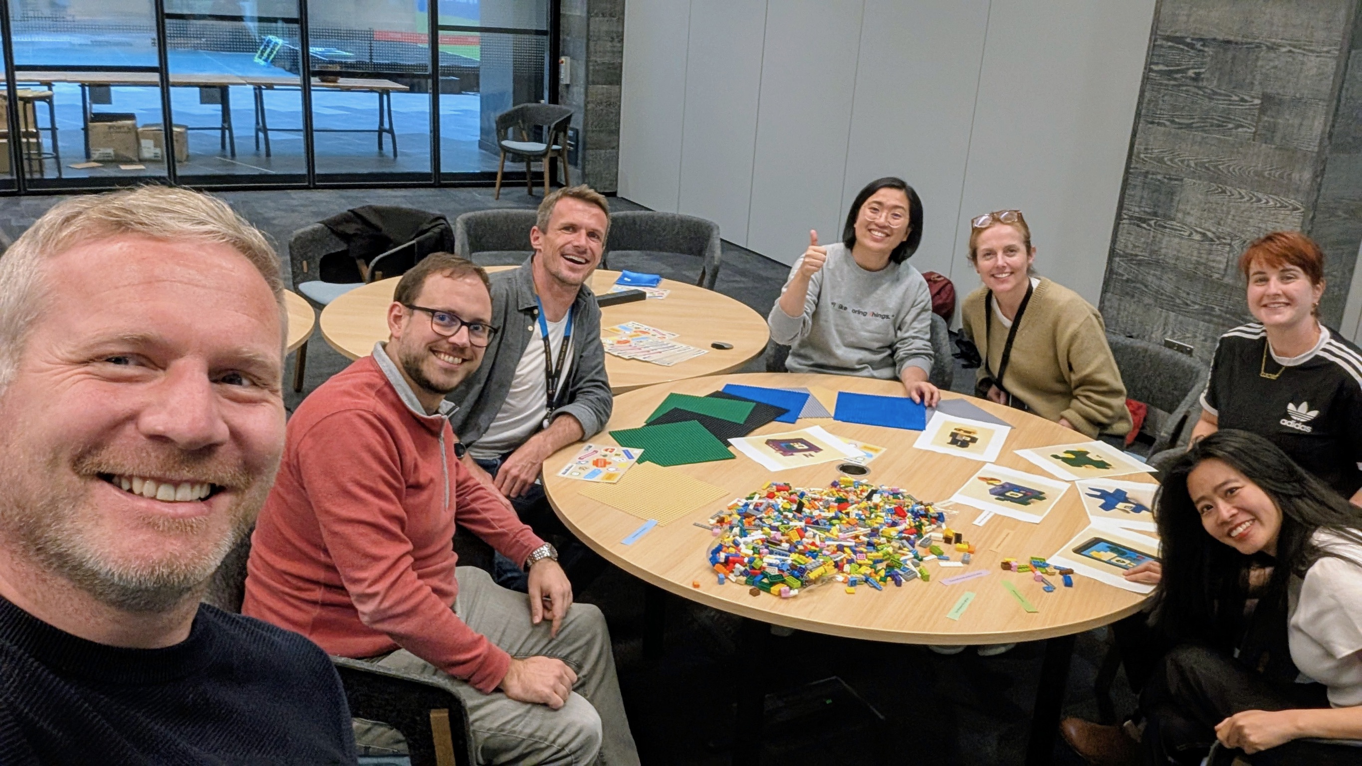 A group of seven people sit around a table covered with LEGO pieces, papers, and cards, smiling at the camera. One person gives a thumbs up. They are in a modern, glass-walled meeting room.
