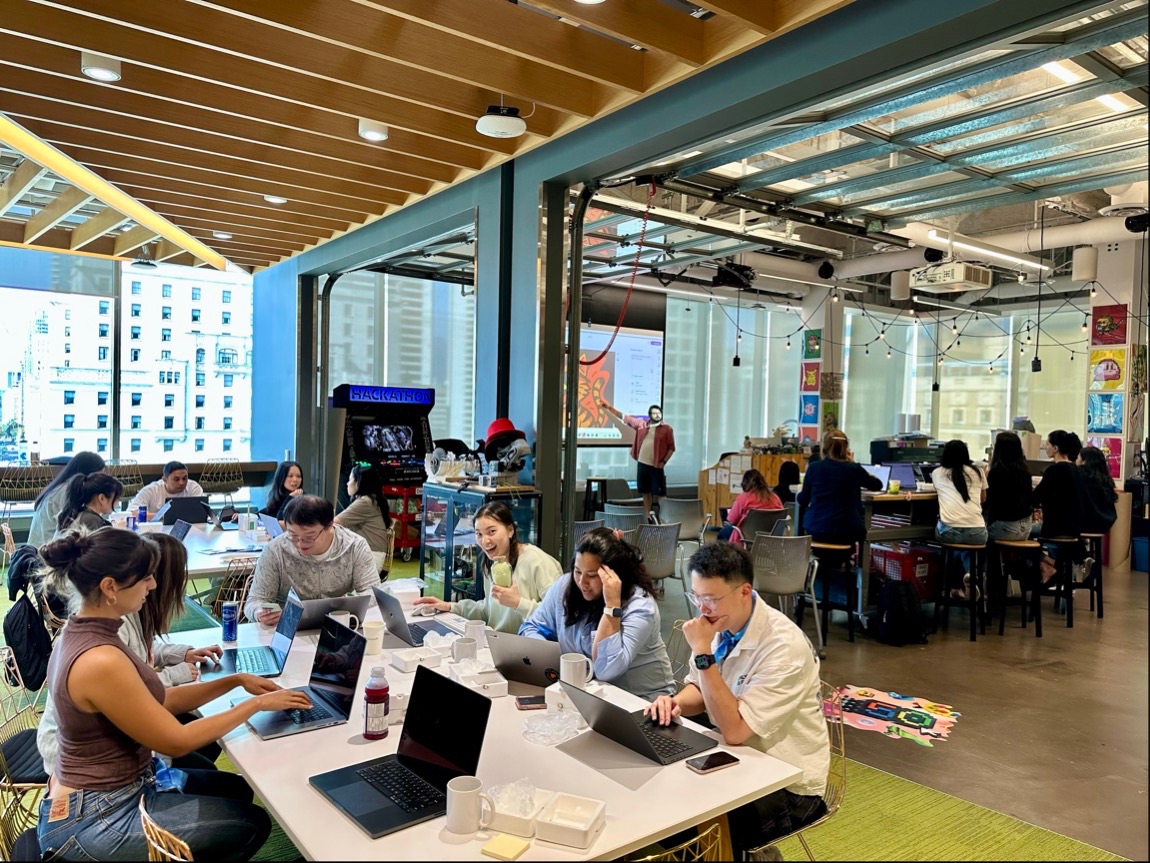 A busy, modern office space with people working on laptops at tables. Some are collaborating in groups, while others focus individually. Large windows let in natural light, and colorful decorations brighten the room.
