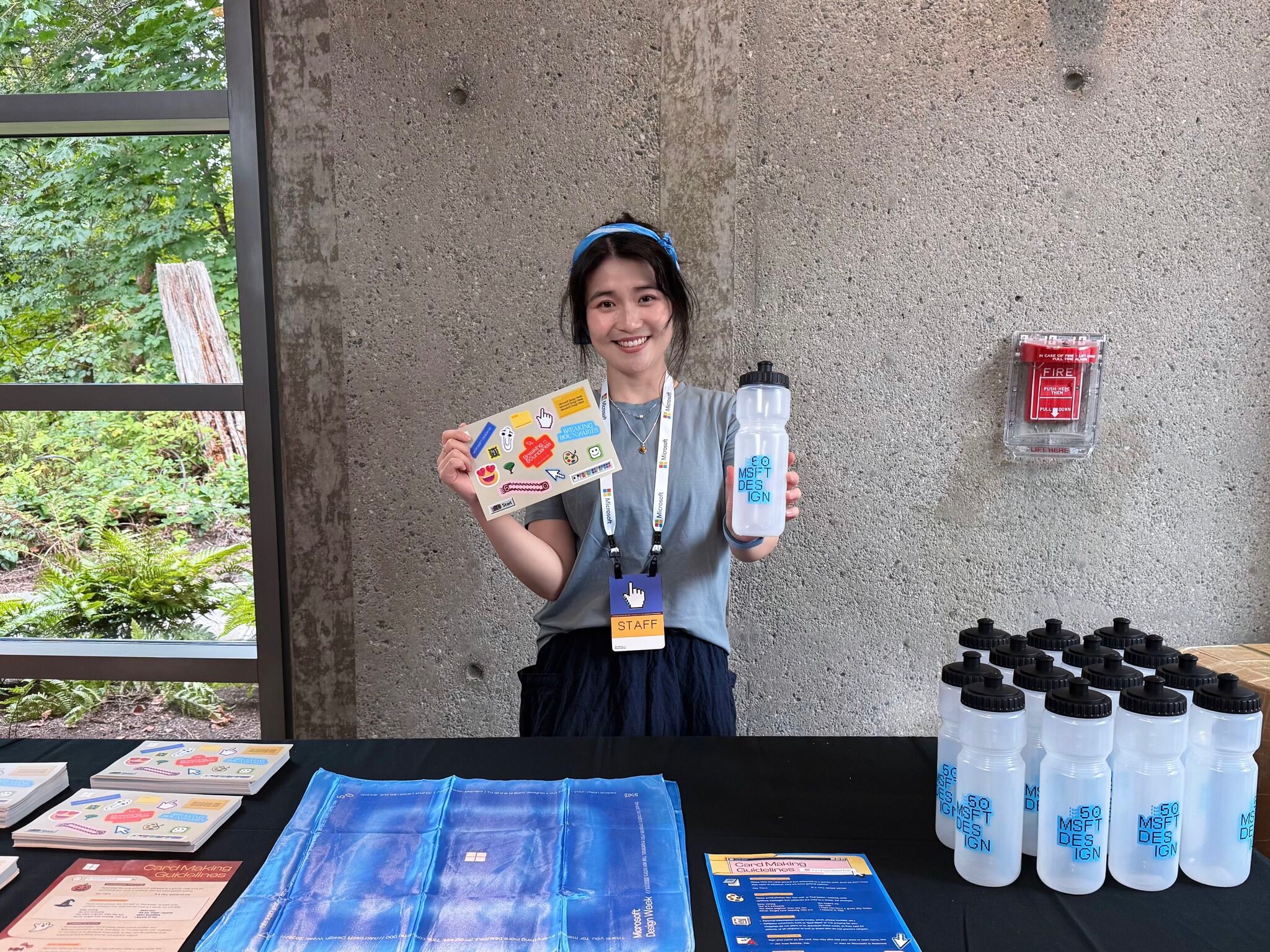 A woman wearing a staff badge stands behind a table with brochures and water bottles. She holds a flyer in one hand and a bottle in the other, smiling. The table is near a window and a gray concrete wall with a fire alarm.