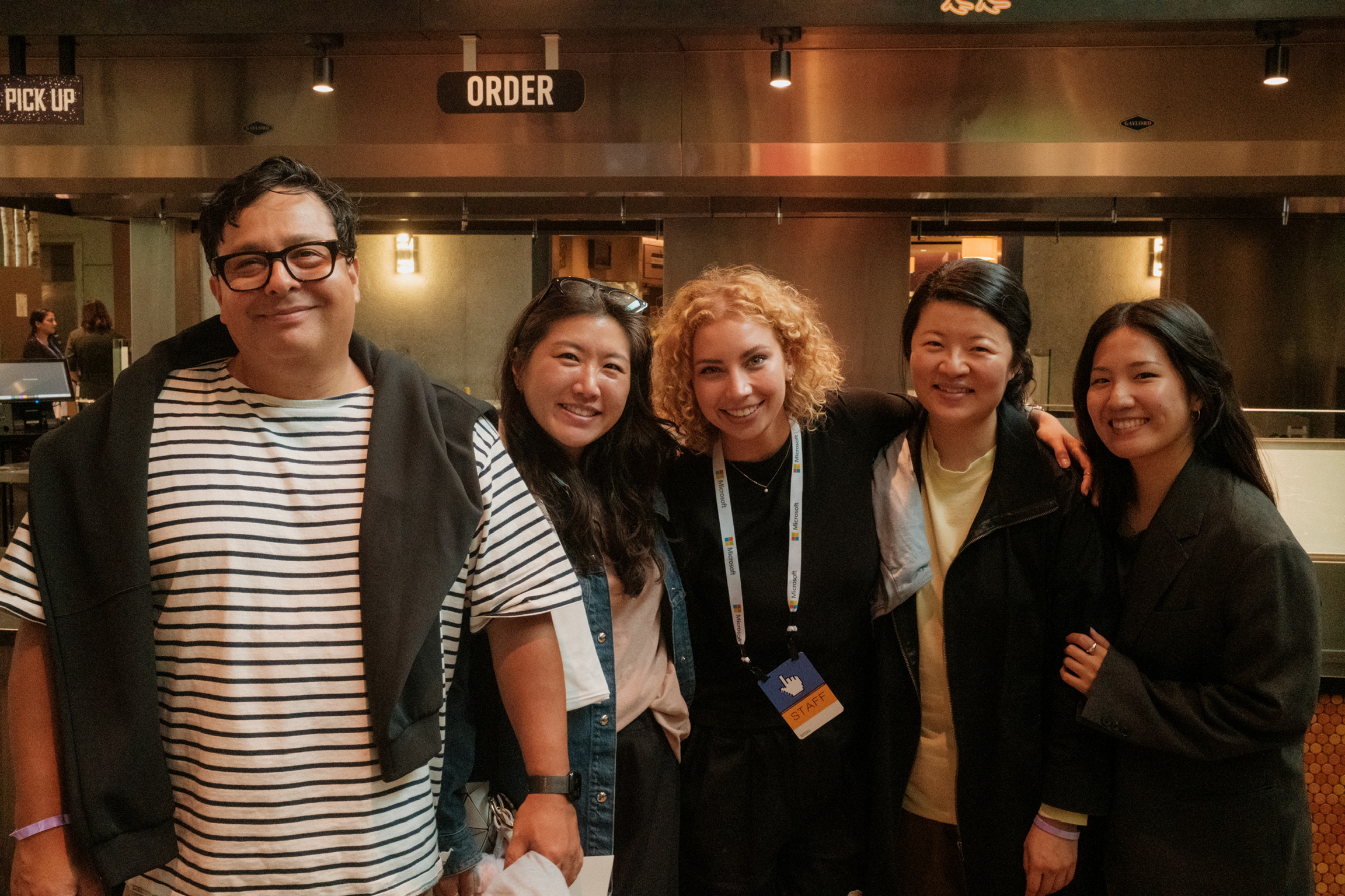 Five people stand close together indoors, smiling at the camera. They appear to be friends or colleagues at an event or restaurant, with an "ORDER" sign visible in the background.