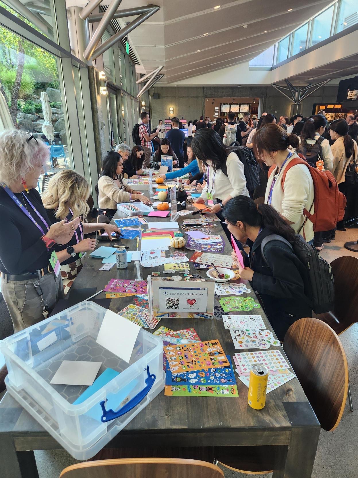 People gather around a large table covered with stickers, paper, and craft supplies, working on arts and crafts projects in a bright, modern indoor space with large windows.