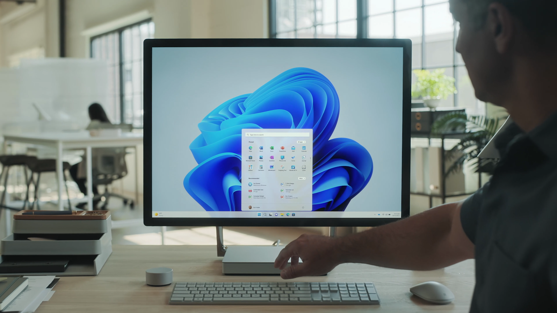 A person uses a desktop computer with a monitor displaying the Windows 11 home screen in a bright, modern office space. The person's hand reaches toward the keyboard.