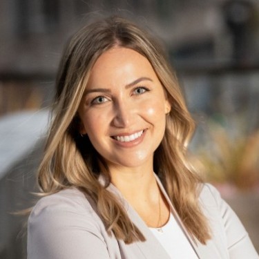 A woman with long, blonde hair smiles at the camera. She is wearing a light-colored blazer over a white top, and the background is softly blurred with outdoor scenery.