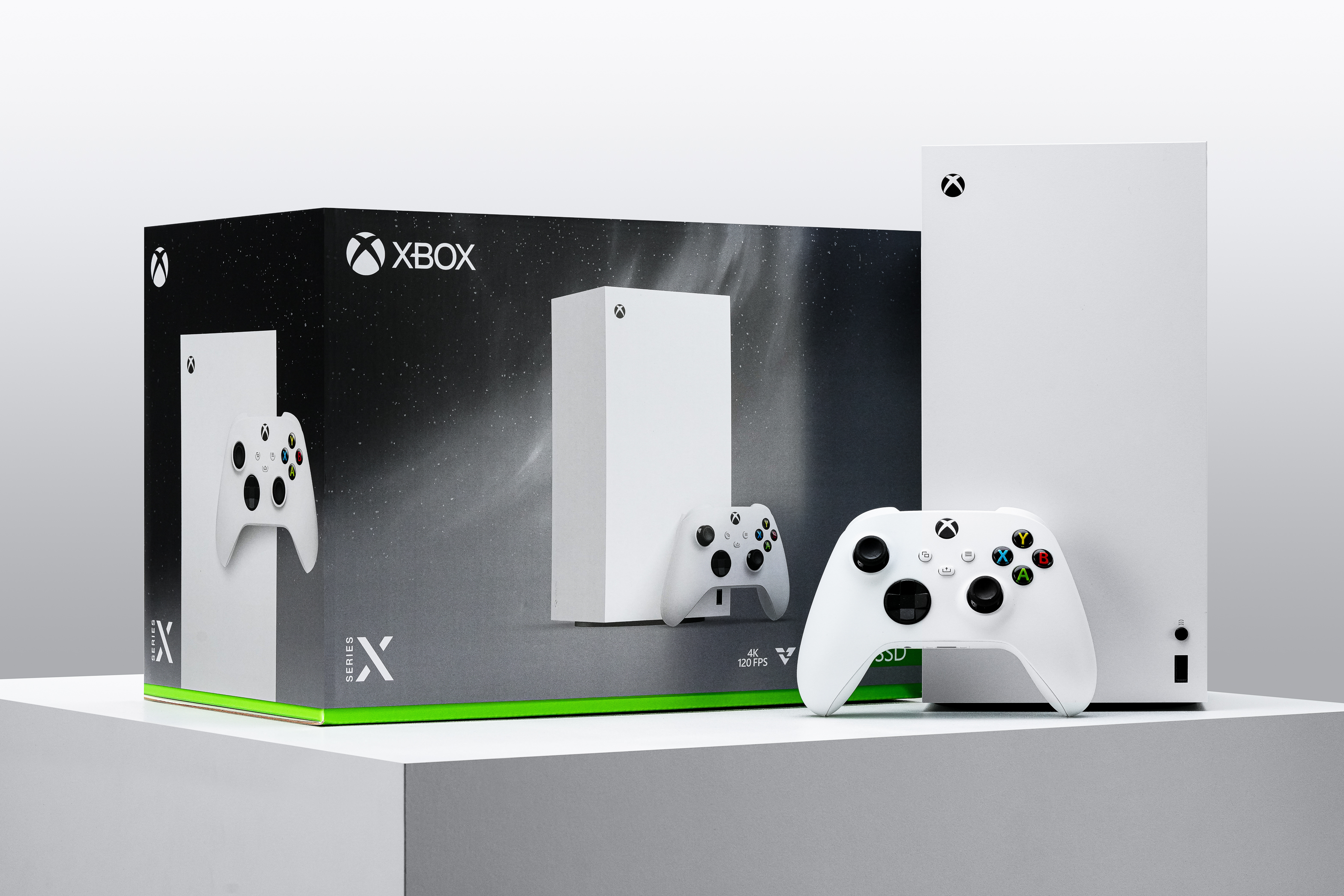 A white Xbox Series X console and controller are displayed on a pedestal next to the console’s retail box, which features product images and branding. The background is minimal and gray.