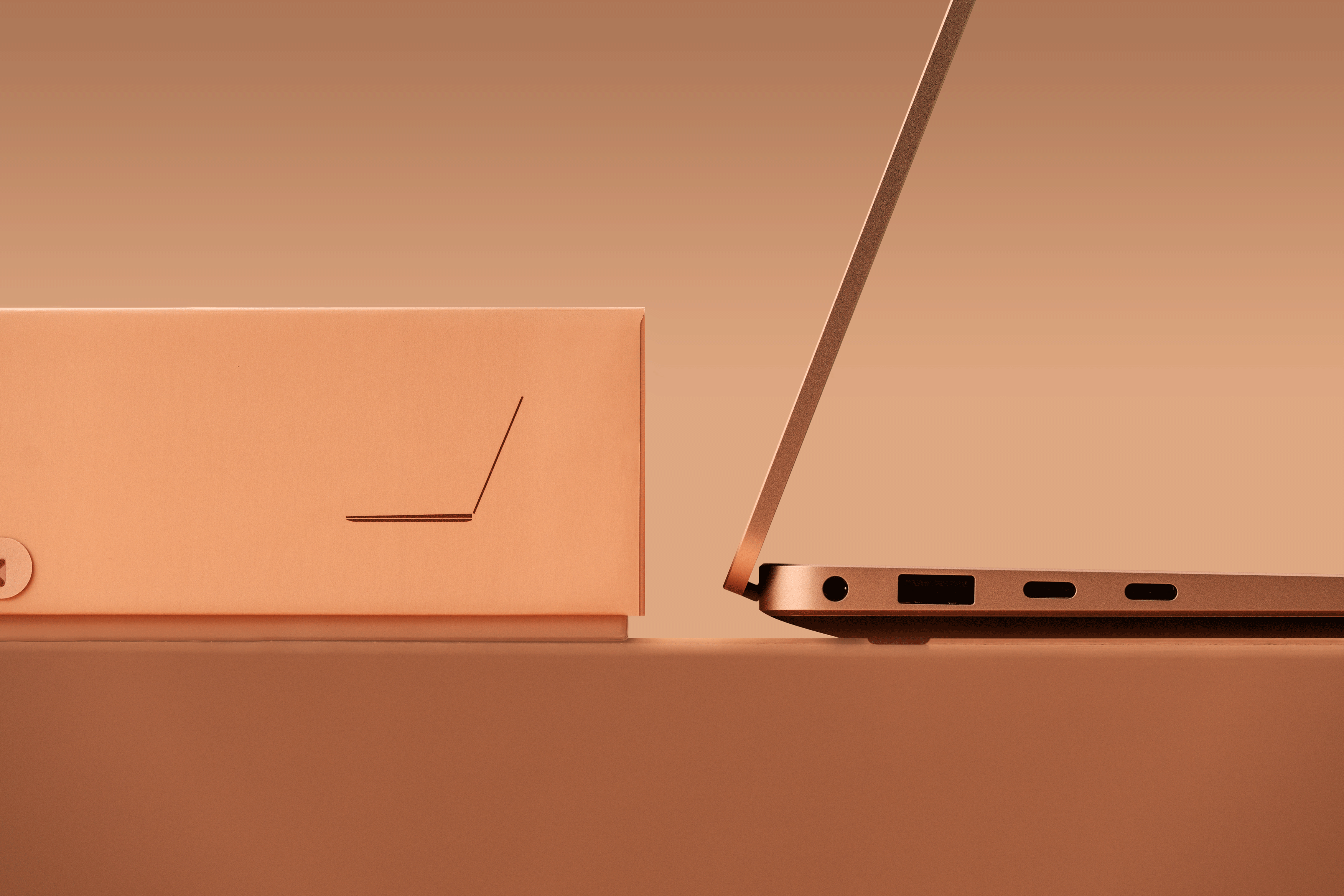 A sleek, partially open laptop sits on a smooth surface next to a rectangular box with a minimalist line design, set against a warm, monochromatic beige background.