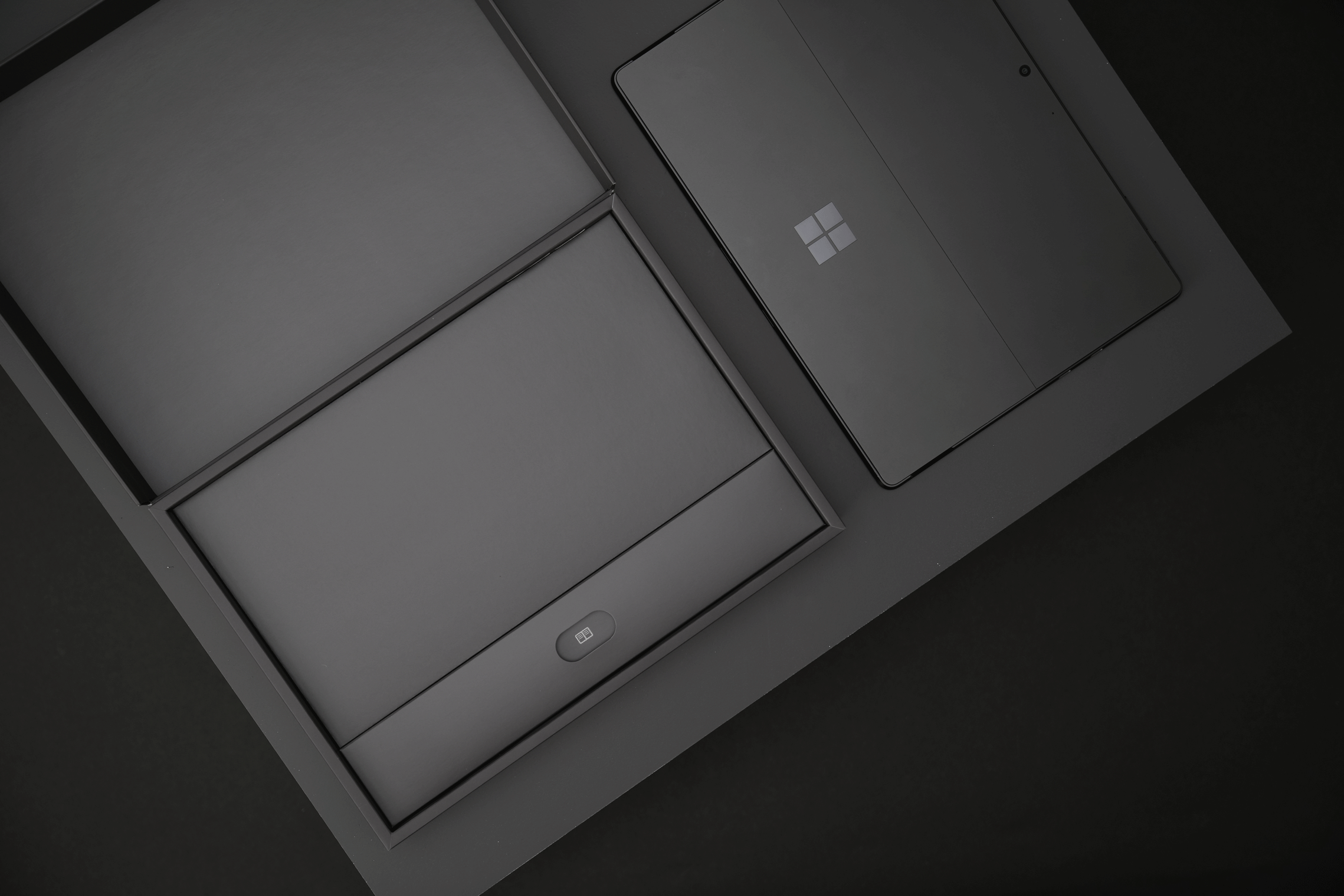 A sleek, dark-colored Microsoft Surface laptop and accessories are neatly arranged in a box with minimalist packaging, viewed from above on a black background.