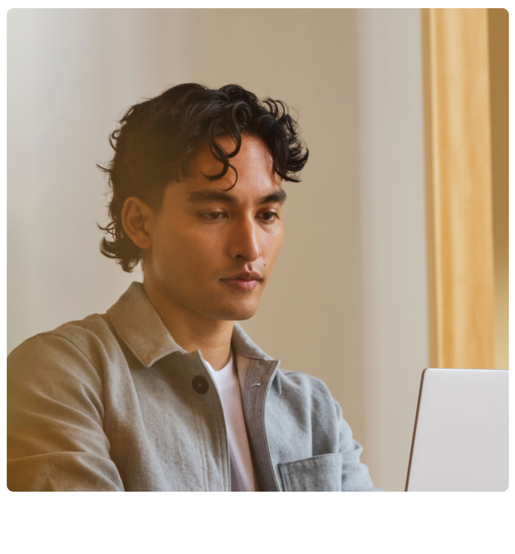 A person with wavy dark hair wearing a light gray jacket looks focused while using a laptop in a bright indoor setting.