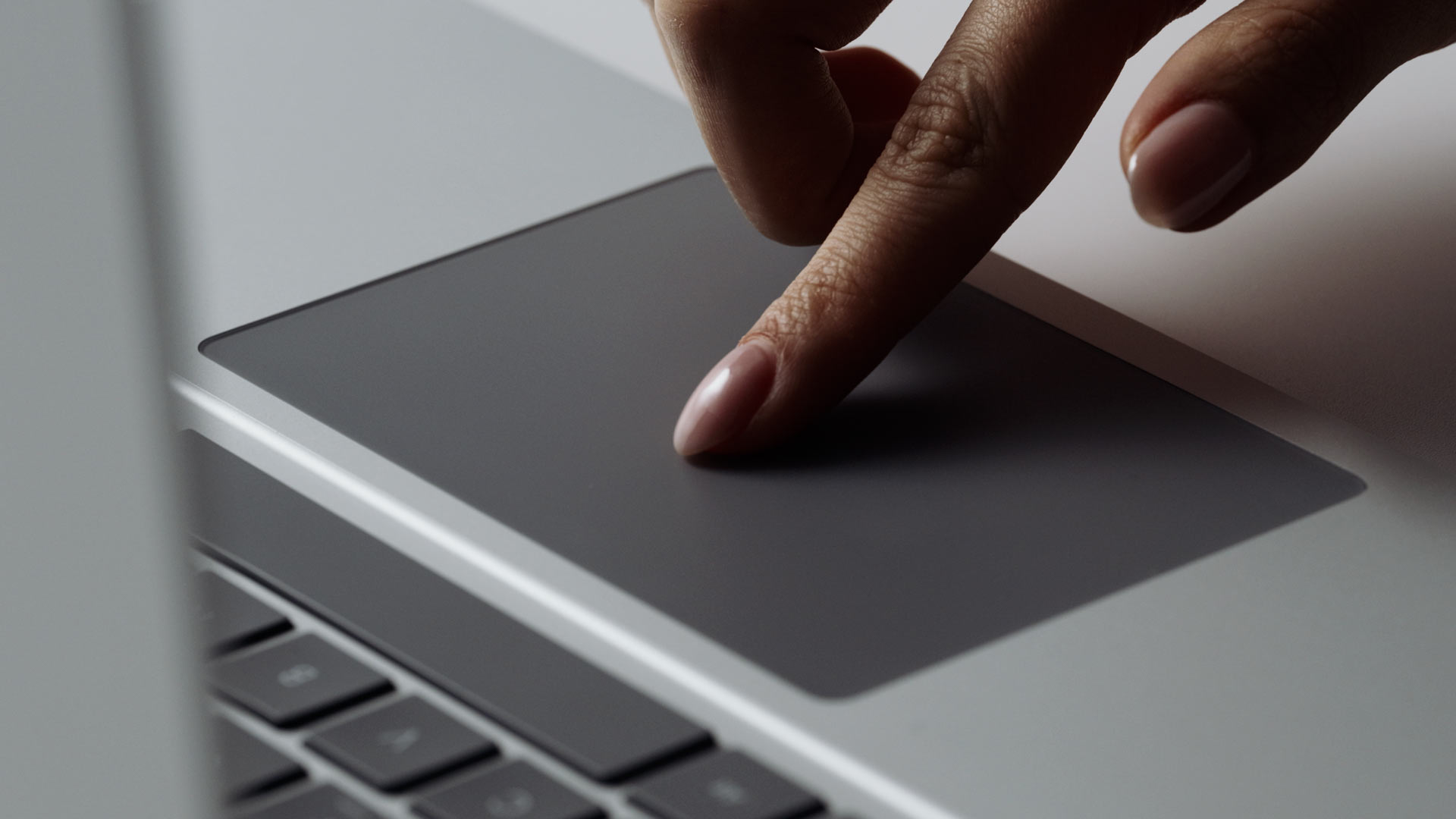A close-up of a person’s hand using the touchpad on a laptop, with their index finger pressed on the trackpad and part of the keyboard visible nearby.