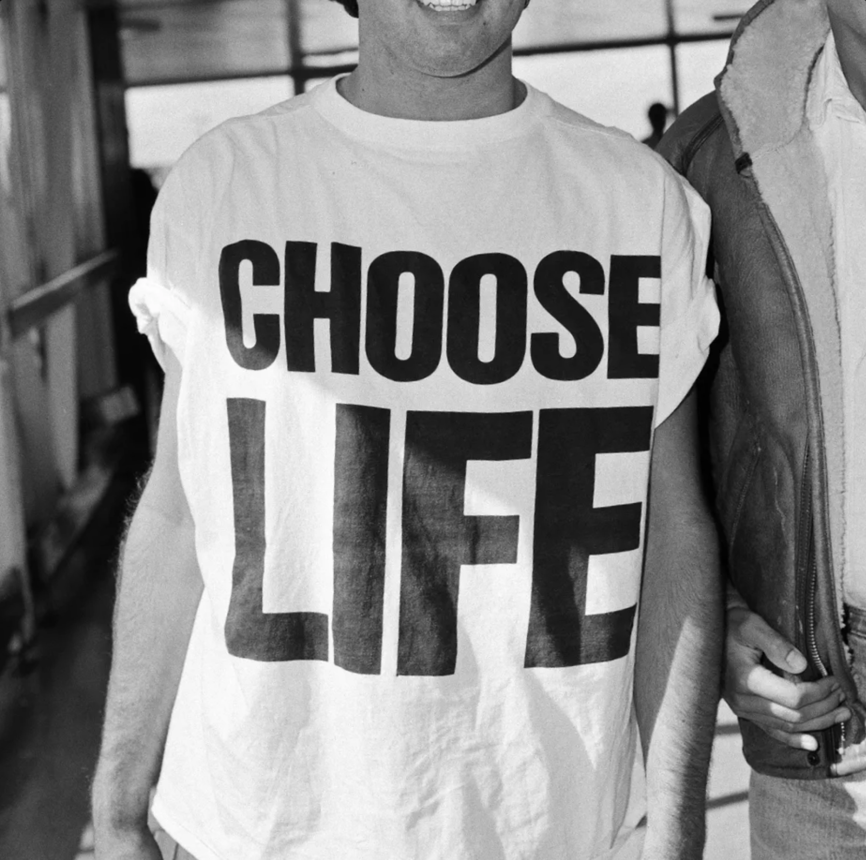 A person wearing a white t-shirt with large black letters that read "CHOOSE LIFE" stands indoors. Their face is partially out of frame, and another person is standing beside them.