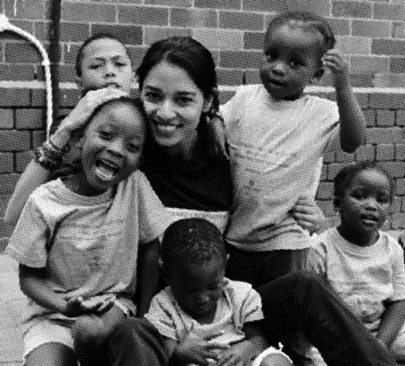A woman sits outside with five smiling children in casual clothes, posing closely together in front of a brick wall.