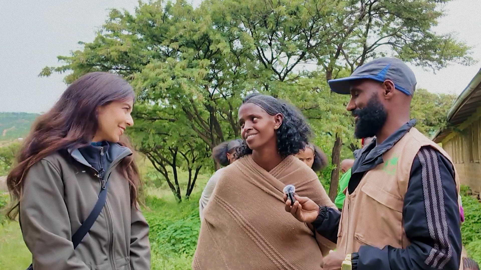 Three people stand outdoors talking and smiling; one man holds a microphone towards a woman wrapped in a brown shawl. Trees and greenery fill the background.