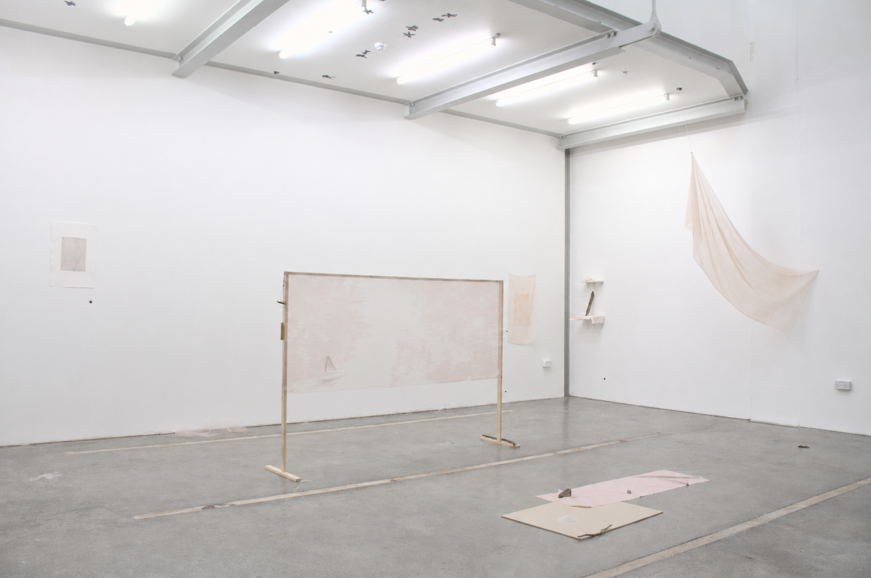 Minimalist art gallery with white walls, soft beige fabric hanging, delicate framed paper, and subtle floor installations. Bright ceiling lights illuminate the sparse, modern space.