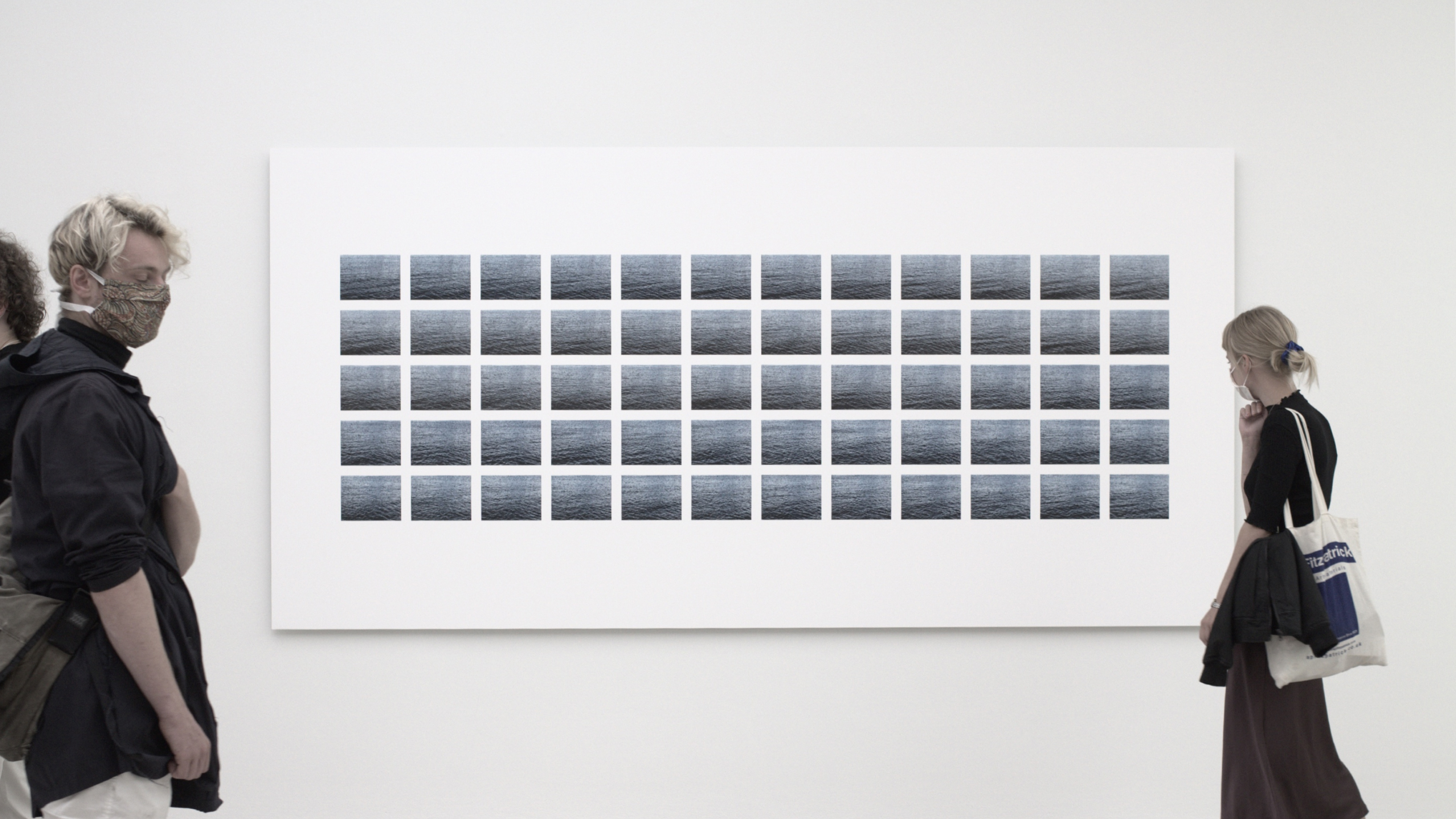 Two people wearing face masks stand on opposite sides of a large white wall displaying a grid of 60 small, identical photographs of rippling water.