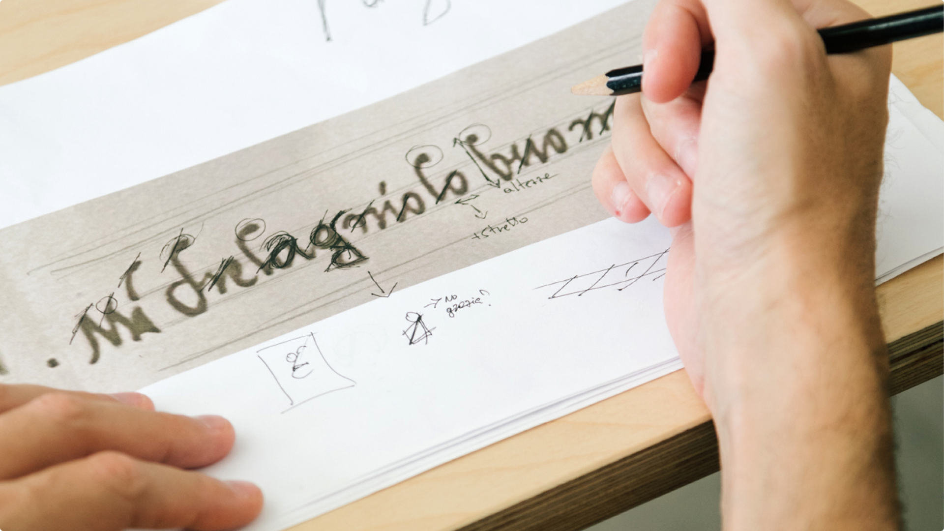 A hand with a pencil sketches corrections and notes over handwritten calligraphy on a strip of paper.