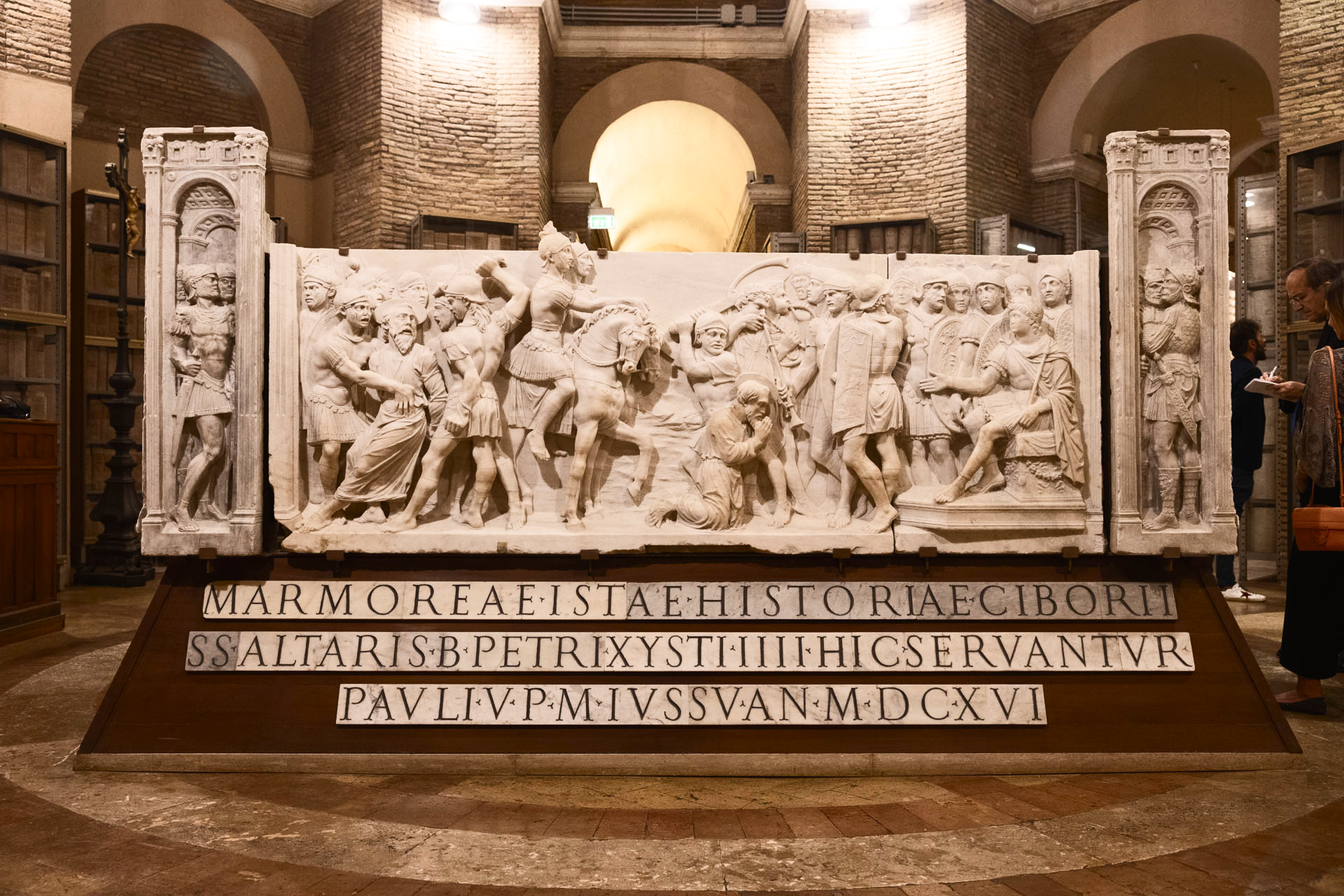 A large marble relief sculpture depicting a battle scene, mounted on a base with a Latin inscription, located inside a vaulted stone chamber.