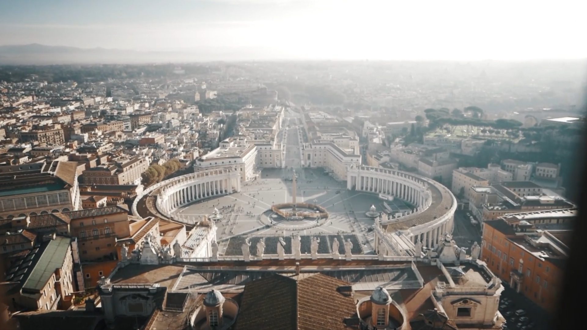 Aerial view of St. Peter’s Square in Vatican City, featuring its iconic elliptical colonnades and central obelisk, surrounded by the rooftops of Rome.